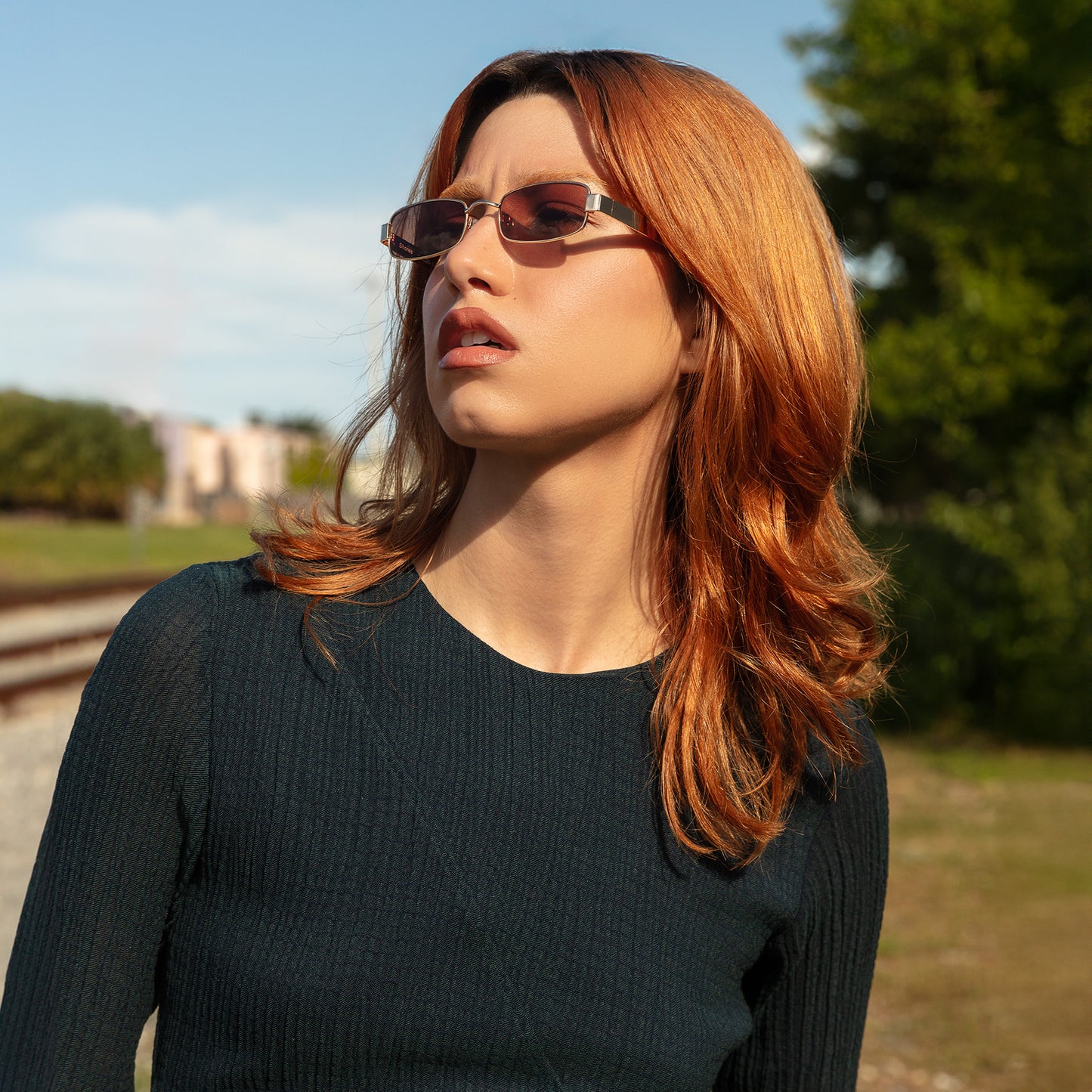 Model wearing Shores Sunglasses in Gold with Brown lenses near train tracks looking to her right